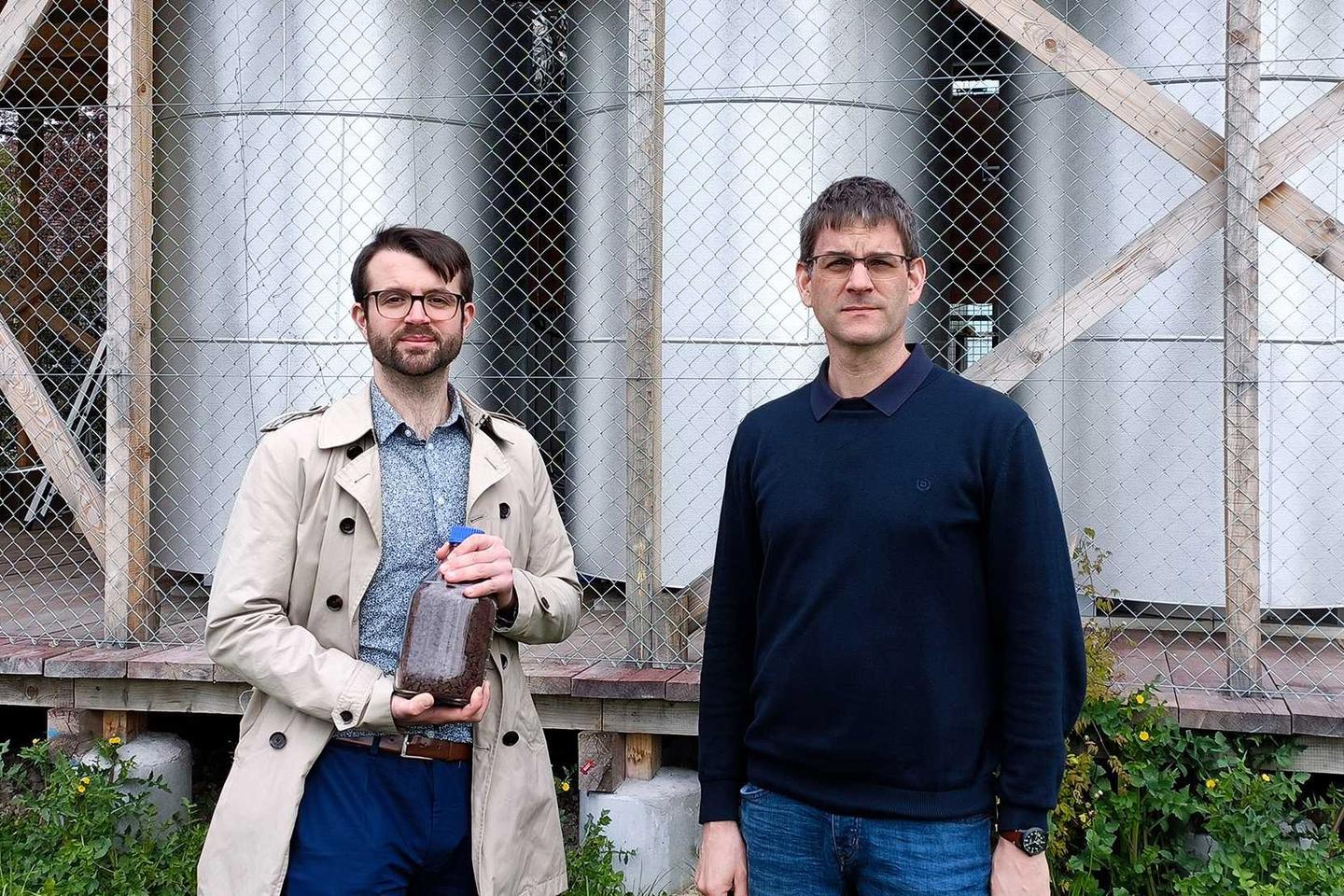 Researchers Samuel Heiniger (left, holding a jar of iron ore) and Professor Wendelin Stark (right) in front of the three iron reactors containing hydrogen